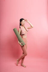 A smiling woman in workout clothes a towel around her neck holds a yoga mat on a pink background