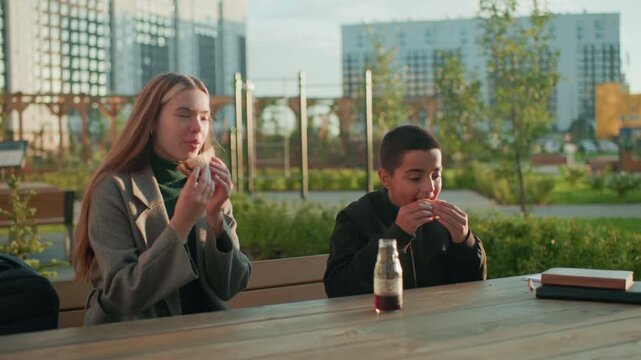 Mum and son sitting outdoors at wooden table in urban park making toast with bread in hand while enjoying snack together, smiling and sharing relaxed family bonding