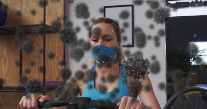 Cycling woman wearing mask on air resistance bike in gym area, showing rolled mats, virus overlay - Powered by Adobe