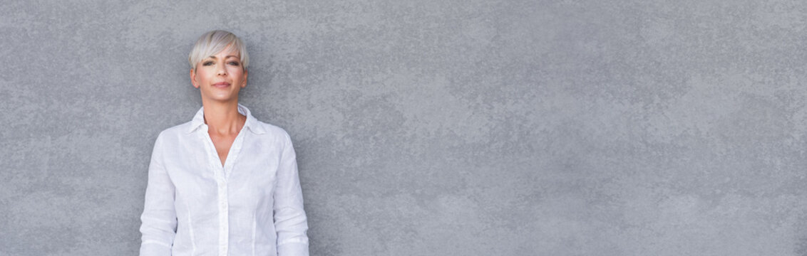 Serious adult woman standing with neutral face expression, against gray background - wall. Pretty middle age woman