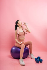 Hydrated and healthy Woman drinking water after workout sitting on fitness ball Pink background