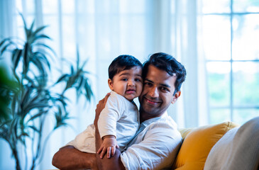 Indian Father and smiling infant boy cuddling on cozy sofa in sunlit modern living space