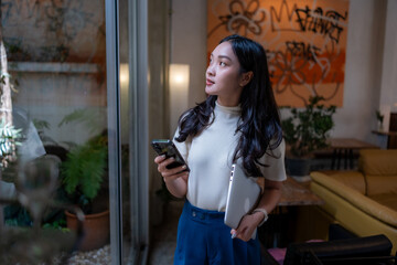 Asian creative woman looking up, holding phone and laptop