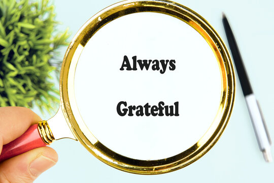 A hand holds a magnifying glass over the words always grateful, symbolizing appreciation and mindfulness in a calm workspace with greenery and a pen nearby