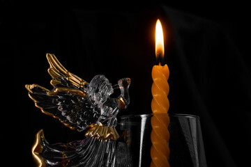 Glass archangel with golden wings blows a trumpet beside a lit spiral candle in a clear glass on black, symbol of mourning, prayer and remembrance for the dead. Photo