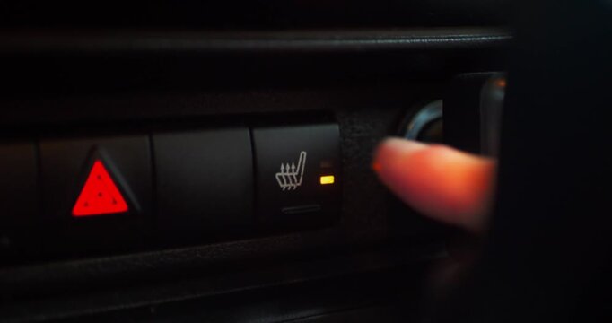 Person presses illuminated heated seat button on car dashboard. Hazard light icon visible. Footage demonstrates modern vehicle interior, driver comfort feature, and automotive technology.​​