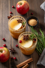 Two glasses of hot apple cider with cinnamon, anise, brown sugar and rosemary on a wooden background