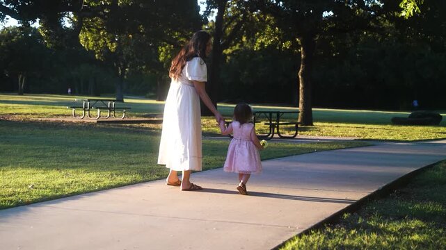 Mother and Small Toddler Daughter Holding Hands and Yellow Flowers While Walking Along Sidewalk in Green Park at Golden Hour of Sunset