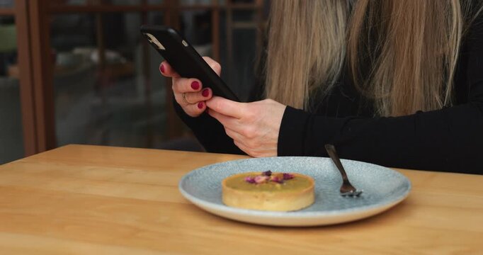 Stylish woman with phone scrolls browsing internet in cafe. Faceless person with long loose hair sits at table with dessert on plate using mobile phone
