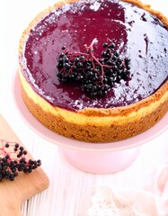 A cheesecake topped with a dark, glossy glaze and decorated with a cluster of dark berries. The cheesecake is presented on a pink cake stand, with a wooden cutting board and berries in the foreground.