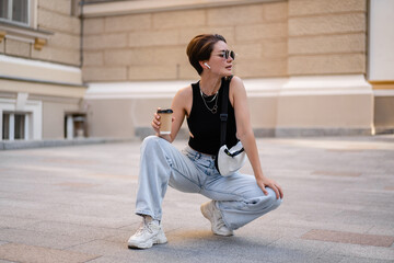 stylish woman with short haircut walking in street with coffee wearing black top, jeans and sunglasses accessories
