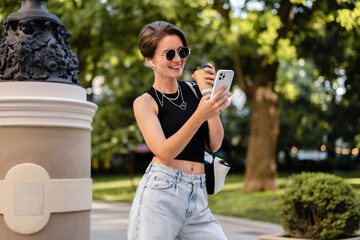 stylish woman with short haircut walking in street with coffee wearing black top, jeans and sunglasses accessories