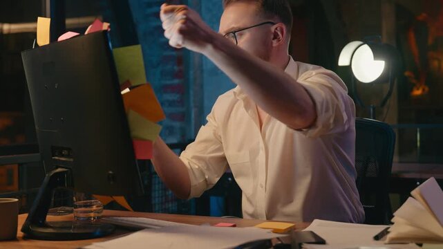 Irritated Caucasian business man at night throwing away stickers from monitor office computer stressed exhausted overworked businessman male guy check throw sticky notes with work tasks holding head