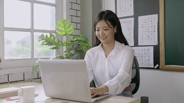 Happy beautiful young Asian female teacher sitting smiling at her desk and using a computer laptop to preparing lessons for teaching her students in the classroom at to school. Back to school concept.