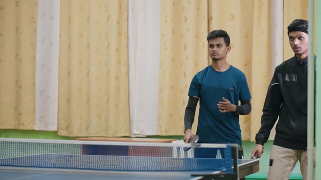 Young teammate in blue shirt with black undershirt competes in indoor table tennis match, focused on ball movement across net, neutral expression with yellow curtain background