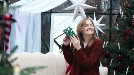 Cheerful woman sitting on a sofa surrounded by Christmas trees and decorations, holding a wrapped green present and smiling happily. Cozy festive atmosphere full of joy and anticipation. Perfect for