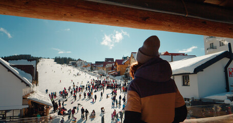 Back view of woman dancing while standing on winter resort balcony looking the tourists. View from the hotel balcony to the snowing ski path resort.