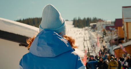 Back view close up shot of woman walking towads the ski path. Skier in blue hat and parka going down resort pathway.
