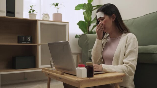 Working from home concept. Young Asian woman employee is symptoms sick and has a headache from the flu use fever cooling gel patch and sitting using laptop computer for working in living room at home.