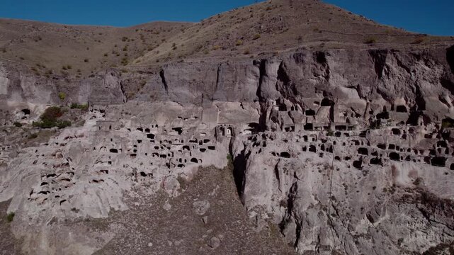 Drone flying over the ancient cave city of Vardzia in Georgia &mdash; a vast monastery and fortress carved into a mountain cliff, illuminated by summer sunlight with birds flying nearby