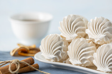 Whipped meringue cookies arranged on a white plate, accompanied by a cup of dark coffee, cinnamon sticks, and dried citrus slices on a textured blue tablecloth