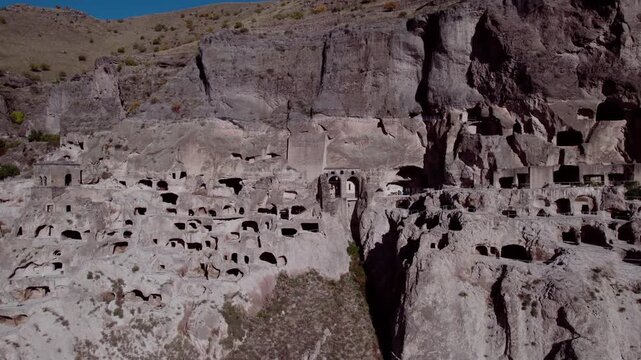 Drone flying over the ancient cave city of Vardzia in Georgia &mdash; a vast monastery and fortress carved into a mountain cliff, illuminated by summer sunlight with birds flying nearby