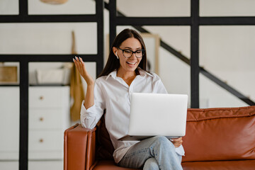 woman sitting on sofa at home relaxed using laptop in white t-shirt and jeans listening to earpods