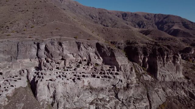Drone flying over the ancient cave city of Vardzia in Georgia &mdash; a vast monastery and fortress carved into a mountain cliff, illuminated by summer sunlight with birds flying nearby