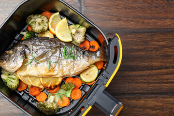 Cooked fish, lemon and vegetables in air fryer basket on wooden table, top view. Space for text