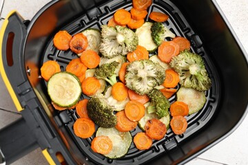Cooked carrots, broccoli and zucchini in air fryer basket on white tiled table, top view