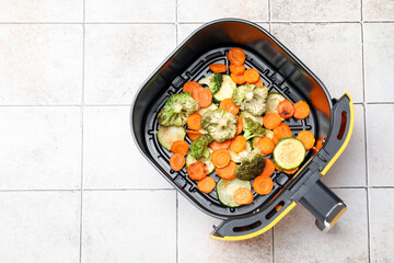 Cooked carrots, broccoli and zucchini in air fryer basket on white tiled table, top view. Space for text