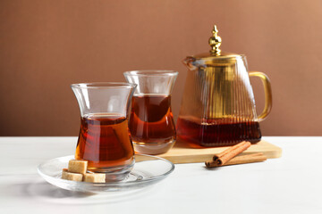 Tasty Turkish tea in glass cups, teapot, brown sugar and cinnamon on white marble table against color background, closeup