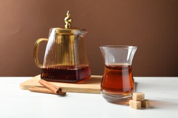 Tasty Turkish tea in glass cup, teapot, brown sugar and cinnamon on white marble table against color background, closeup