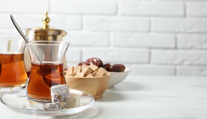 Tasty Turkish tea in glass cups, brown sugar and sweets on white marble table against brick wall, closeup. Space for text