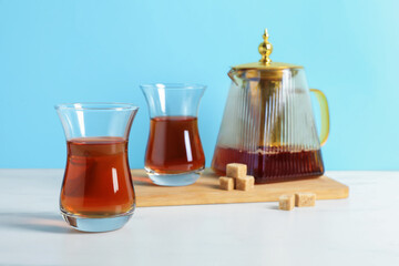 Tasty Turkish tea in glass cups, teapot and brown sugar on white marble table against light blue background