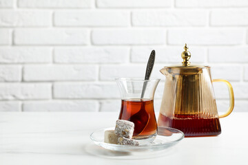 Tasty Turkish tea in glass cup, teapot and sweets on white marble table against brick wall, closeup. Space for text