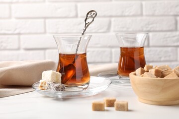 Tasty Turkish tea in glass cups, brown sugar and sweets on white marble table against brick wall, closeup