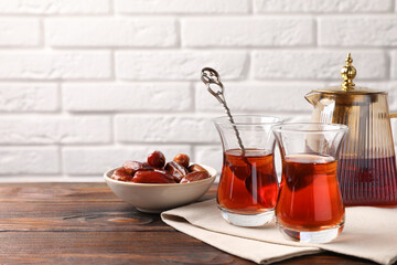 Tasty Turkish tea in glass cups, dates and teapot on wooden table against white brick wall, closeup. Space for text