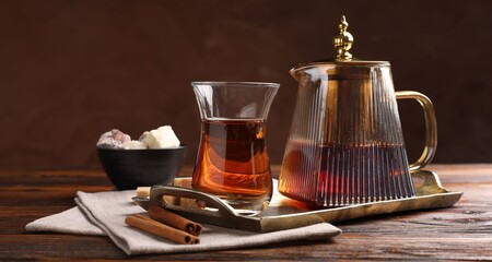 Tasty Turkish tea in glass cup, brown sugar, cinnamon, sweets and teapot on wooden table against color background, closeup