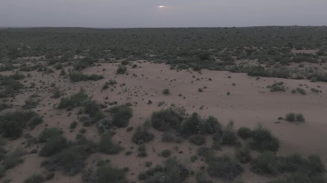 Aerial view of Khuri Sand Dunes near Jaisalmer, Rajasthan, with the sun setting behind a cloudy sky, revealing golden dunes merging with green desert vegetation like Khejri, Rohida, and Babool trees