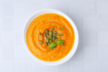 Delicious pumpkin soup with seeds and basil in bowl on white tiled table, top view