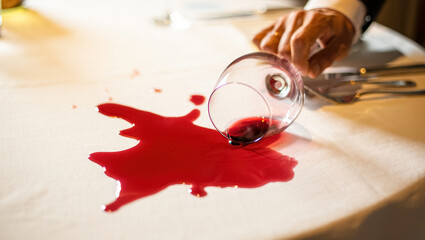 Close-up of a hand next to an overturned glass with spilled red wine spreading across a white tablecloth in warm evening restaurant light