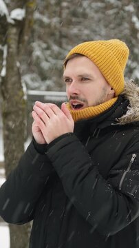 Caucasian man in warm clothing rubs hands and warms them with breath during snowfall outside. Vertical video of portrait of person waiting in snowy city.