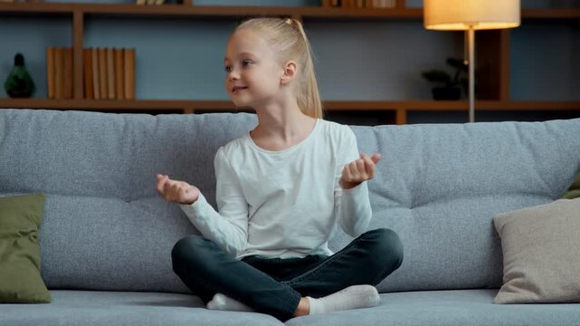 Caucasian little child girl dancing carefree sitting on couch at home domestic joy kid in lotus pose snap fingers dance to listening energetic music cheerful satisfied daughter moving happy childhood