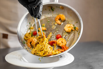 Chef plating creamy shrimp fettuccine with cherry tomatoes and herbs in a professional kitchen. Close-up of hands in gloves using tongs for elegant food presentation.