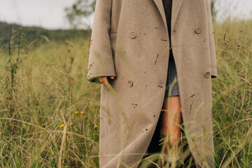 Close-up of an oversized beige coat on an unrecognizable woman standing in a wild meadow.
