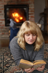 A woman reads a book lying on the floor near the fireplace enjoying a peaceful evening.