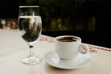 White porcelain cup with strong Turkish black coffee or americano and glass of water served on an outdoor cafe table in natural light.