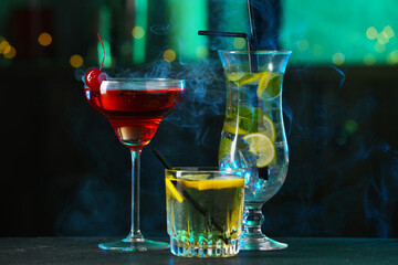 Glasses of tasty cocktails on dark table against blurred background with smoke, bokeh effect