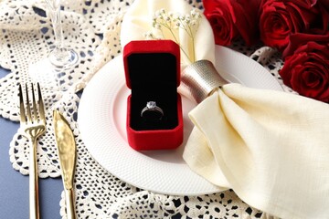 Beautiful table setting for romantic dinner, box with engagement ring and roses on blue background, closeup
